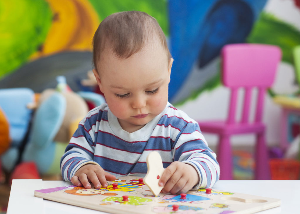Small toddler or a baby child playing with puzzle shapes on a low table in a colorful children room in a nursery or preschool.